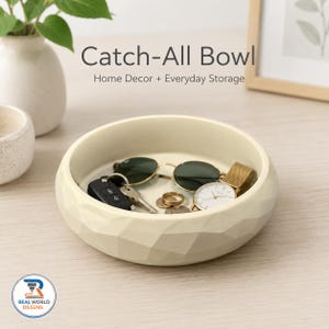 May include: A cream-colored, round catch-all bowl with a faceted, geometric exterior texture. The bowl contains a pair of sunglasses, a black car key fob, a gold-tone wristwatch, and several small metal coins resting inside the basin.