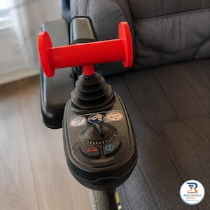 May include: A black electric wheelchair joystick controller featuring a bright red T-shaped handle attachment. The control panel includes a central dial, a horn button, a power button, and three circular safety warning stickers on the top surface.