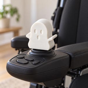 Puede incluir: Un accesorio para joystick en forma de fantasma blanco con dos ojos ovalados y una boca ovalada, diseñado para encajar en la palanca de control negra de una silla de ruedas eléctrica. La figura tiene brazos pequeños extendidos y acabado mate.