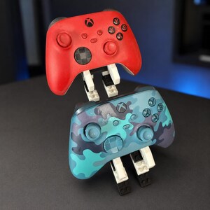 May include: A red wireless video game controller and a blue camouflage patterned wireless video game controller, both mounted on a white and black plastic building block stand that holds the two controllers in a stacked vertical position.