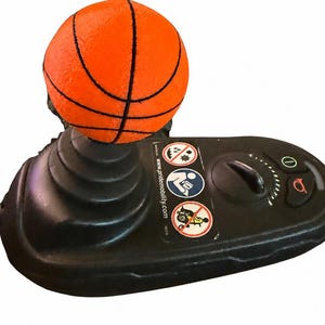 May include: A black mobility scooter joystick controller topped with an orange basketball-shaped handle. The base features a speed dial, a horn button, a power button, and three circular safety warning labels including the text www.pridemobility.com.
