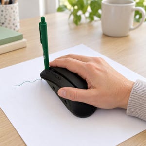 May include: A black ergonomic pen holder attachment for a computer mouse, featuring a vertical slot that holds a green ballpoint pen. The device is positioned on a white sheet of paper, allowing the pen to make contact with the surface for drawing.