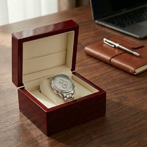 May include: A silver wristwatch with a stainless steel strap sits in an open, dark red wooden watch box. The box has a cream-coloured interior and is on a wooden surface. A leather notebook and pen are visible in the background.