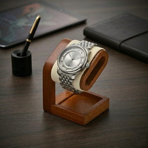 May include: A silver wristwatch with a stainless steel band is displayed on a wooden watch stand with a cream-colored cushion. The watch has a silver face and is set against a dark wooden surface. A pen and tablet are in the background.