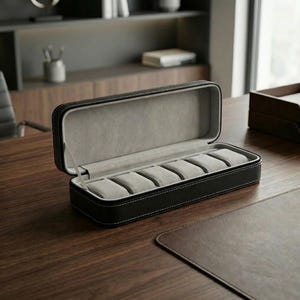 Peut inclure: Étui à montres en cuir noir avec six compartiments rembourrés gris. Le boîtier rectangulaire est ouvert, révélant l'intérieur. Des coutures blanches ornent les bords. L'étui est posé sur un bureau en bois.