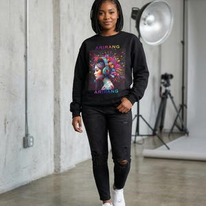 Könnte beinhalten: Schwarzes Sweatshirt mit einem farbenfrohen Grafik einer Frau mit Kopfhörern, mit dem Text "ARIRANG" über und unter dem Bild. Die Frau trägt eine schwarze Jeans und weiße Turnschuhe.