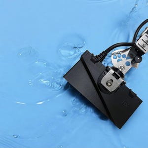 Può includere: Un dispositivo nero con un componente metallico argentato e un cavo nero, parzialmente immerso in acqua blu. Il dispositivo ha una forma rettangolare con le iniziali "BS" visibili. Il componente argentato ha un design a impronta di zampa.