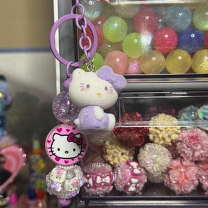Peut inclure: Un porte-clés violet avec deux breloques Hello Kitty. L'une est une figurine Hello Kitty assise avec un nœud violet. L'autre est une breloque Hello Kitty rose et blanche avec une base décorative. Le porte-clés est attaché à une vitrine transparente.