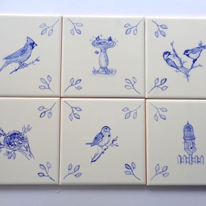 May include: Six square ceramic tiles with blue bird illustrations. Each tile features a different bird or bird-related design, including a cardinal, bird bath, and birdhouse. The tiles are arranged in a 2x3 grid, with blue leaf accents.