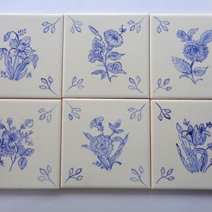 May include: Six square ceramic tiles with hand-painted blue floral designs on a cream background. Each tile features a different flower, with small blue leaf accents in the corners. A decorative set.