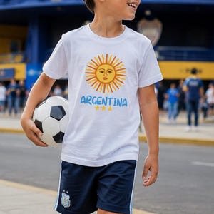 Puede incluir: Una camiseta blanca con un gráfico de sol amarillo con cara y el texto azul ARGENTINA sobre tres estrellas amarillas, combinada con pantalones cortos deportivos azul marino con rayas laterales azul claro y un pequeño emblema, calcetines y zapatillas.