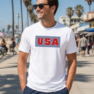 May include: White t-shirt with the word "USA" in red, outlined with blue stripes. The shirt is short-sleeved and worn by a person wearing sunglasses. The background shows a sunny outdoor scene with palm trees and people.