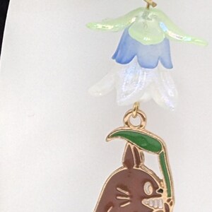 Puede incluir: Pendientes colgantes dorados. La parte superior presenta un lazo dorado y un grupo de abalorios de flores azules y blancas transl&uacute;cidas. El dije inferior es un personaje Totoro marr&oacute;n que sostiene una hoja verde.