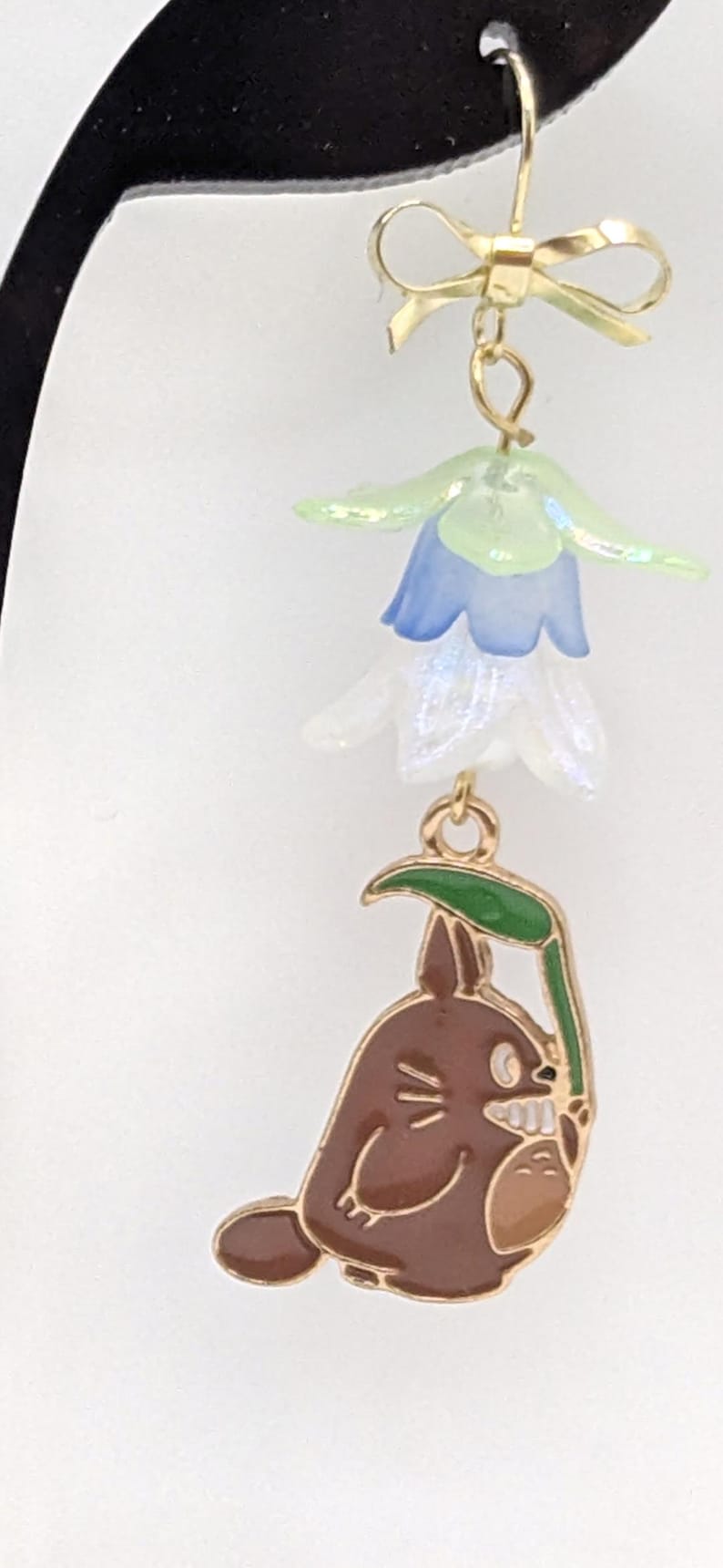 Puede incluir: Pendientes colgantes dorados. La parte superior presenta un lazo dorado y un grupo de abalorios de flores azules y blancas transl&uacute;cidas. El dije inferior es un personaje Totoro marr&oacute;n que sostiene una hoja verde.