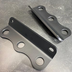 May include: Two black metal brackets with rounded edges and three circular holes each. One bracket is positioned horizontally, while the other is angled upwards. The brackets are on a gray surface.