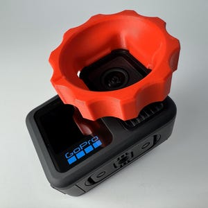 Puede incluir: Una cámara GoPro negra con un accesorio naranja brillante adjunto. La GoPro tiene la palabra "GoPro" en azul en la parte delantera. El accesorio naranja tiene forma circular con un borde estriado.