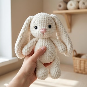 May include: A handmade, off-white crochet bunny with long floppy ears, black eyes, and a pink nose. The plush toy is soft and textured, perfect for a child's comfort. The bunny is held in a hand, showcasing its size and detail.