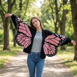 May include: Black crochet cardigan with butterfly wing design. The wings are pink with white and black accents. The cardigan is open-front and has long sleeves. The person in the image is wearing blue jeans and a gray shirt.