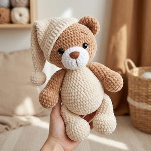 Puede incluir: Un oso de peluche de ganchillo hecho a mano en tonos marrón y crema, con un gorro de dormir a juego con un pompón. El oso tiene ojos de seguridad negros y un hocico blanco. El juguete blando está diseñado para la comodidad.