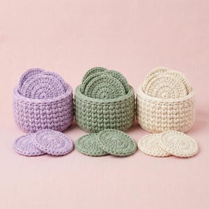 May include: Three sets of crochet coasters in lavender, sage green, and cream, each in a matching basket. The coasters are round with a spiral design. The baskets are cylindrical with a textured stitch.