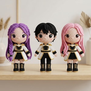 May include: Three handmade crochet dolls with different hair colors: purple, black, and pink. Each doll wears a white and black outfit with gold accents and black boots. The dolls are displayed on a wooden surface.