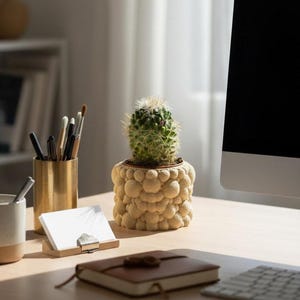 以下が含まれることがあります： ベージュのテクスチャ加工の鉢に入った小さな緑色のサボテンが木製の机の上にあります。 その他のデスクアクセサリーには、ペンとブラシが入ったゴールドのペン立て、メモ帳、茶色の革張りのジャーナルが含まれます。 コンピューターモニターが背景にあります。