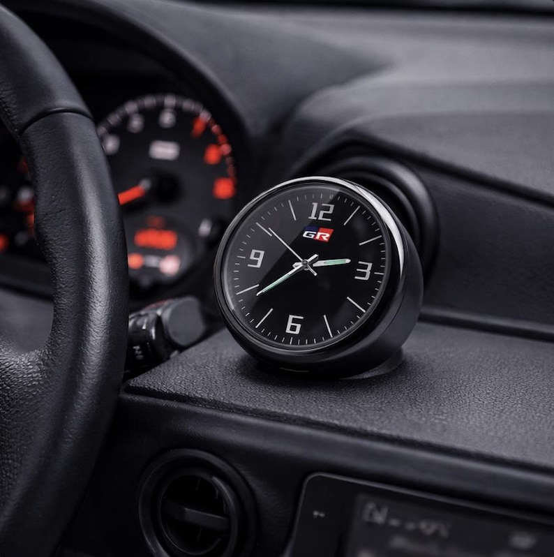 K&ouml;nnte beinhalten: Eine schwarze analoge Uhr mit wei&szlig;en Ziffern und Zeigern, mit den Buchstaben "GR" in Rot und Wei&szlig;. Die Uhr ist auf dem Armaturenbrett eines Autos montiert, mit einem Lenkrad und Anzeigen im Hintergrund.