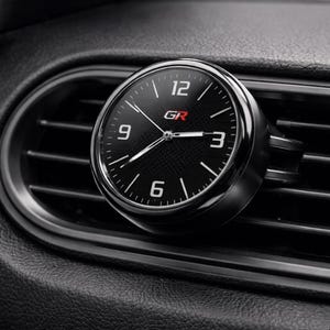 K&ouml;nnte beinhalten: Eine schwarze und silberne analoge Uhr mit den Buchstaben "GR" in Rot. Die Uhr ist an einer L&uuml;ftungs&ouml;ffnung eines Autos befestigt. Das Zifferblatt hat wei&szlig;e Ziffern und Zeiger sowie ein Karbonfasermuster.