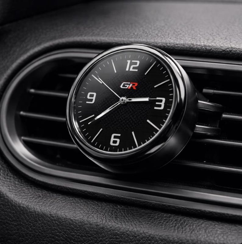 K&ouml;nnte beinhalten: Eine schwarze und silberne analoge Uhr mit den Buchstaben "GR" in Rot. Die Uhr ist an einer L&uuml;ftungs&ouml;ffnung eines Autos befestigt. Das Zifferblatt hat wei&szlig;e Ziffern und Zeiger sowie ein Karbonfasermuster.