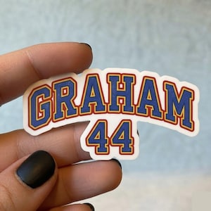 May include: A die-cut sticker featuring the name GRAHAM in a bold, arched varsity-style font, with the number 44 positioned directly below. The text is colored in blue with a thick red outline and a thin white border on a white background.