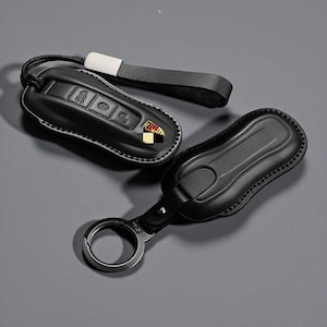 Puede incluir: Dos fundas negras de cuero para llaveros con costuras blancas. Una funda tiene una correa para la muñeca y el logotipo de Porsche. La otra tiene un llavero de metal. Los llaveros tienen botones en relieve.