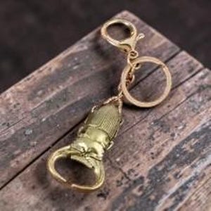 Op de afbeelding: Een goudkleurige metalen sleutelhanger met een gedetailleerde vliegend hert-bedel. De bedel is bevestigd aan een ronde sleutelring en een karabijnsluiting. De kever heeft opvallende kaken en een textuur op de dekschilden van metaal.
