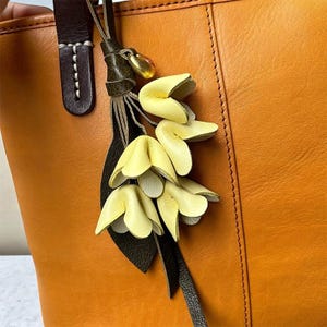 Könnte beinhalten: Nahaufnahme einer orangefarbenen Ledertasche mit einem dekorativen gelben Blumenanhänger. Der Anhänger zeigt drei gelbe Blumen mit weißen Unterseiten, ein dunkelgrünes Blatt und einen goldfarbenen Akzent. Die Tasche hat einen braunen Ledergriff.