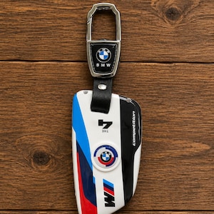 Peut inclure: Un étui pour clé de voiture BMW arborant un design blanc, noir, bleu et rouge avec les inscriptions M, Competition et The 4. L'accessoire comprend une lanière en cuir noir et un clip en métal argenté avec un petit emblème BMW circulaire.