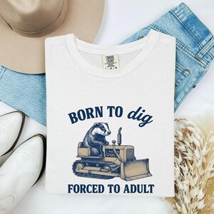 May include: A white t-shirt featuring a dark blue graphic of a badger operating a bulldozer. The text above the badger reads BORN TO dig and the text below reads FORCED TO ADULT. The shirt has a Comfort Colors brand label visible at the inner neckline.