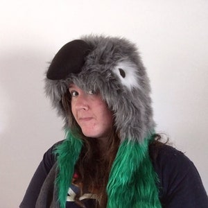 May include: A gray and black bird-themed hat with a black beak and white eye detail. The hat has a fluffy texture and is paired with a green faux fur scarf. The hat is designed to resemble a bird.
