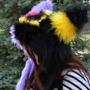May include: A black, yellow, and purple furry hooded hat with a long tail. The hat is made of soft, plush fabric and has a cute, animal-like design.