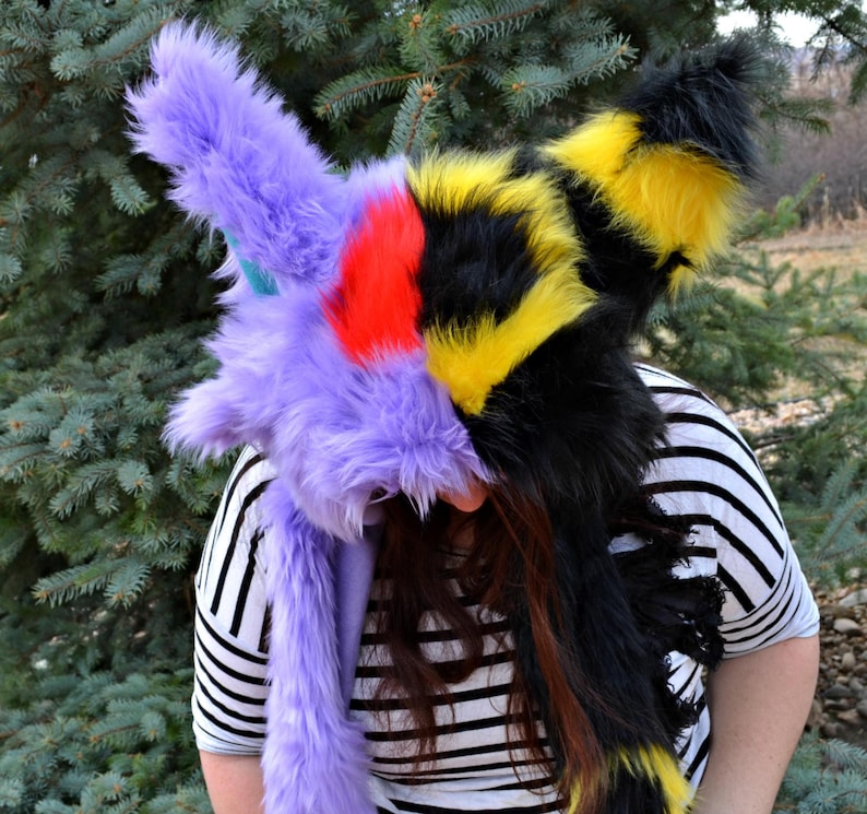May include: A purple and black furry hooded garment with yellow and red accents. The hood is pulled over the head of the person wearing it.