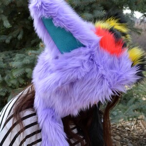 May include: A purple furry hat with long ears and a black, yellow, and red tail. The hat is worn by a person with long brown hair.