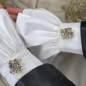 Peut inclure: Gros plan sur des poignets de chemise blancs à volants avec des détails de boutons de manchette ornés d'or et de cristaux. Les poignets sont associés à une veste en cuir noir, créant un contraste de textures et de couleurs. L'esthétique générale est élégante et sophistiquée.