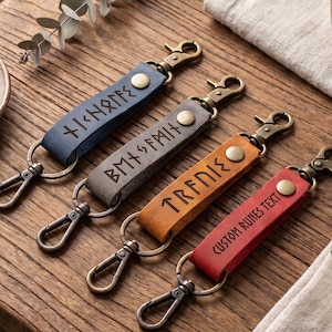 May include: Four leather keychains in blue, gray, tan, and red, each featuring a brass snap button and a metal lobster clasp. The blue, gray, and tan straps are embossed with runic characters, while the red strap displays the text CUSTOM RUNES TEXT.