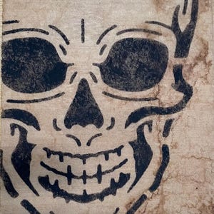 May include: A rectangular piece of aged, stained paper with rough, torn edges featuring a bold, black ink illustration of a human skull. The skull design includes deep eye sockets, a triangular nose cavity, and a detailed jaw with visible teeth.
