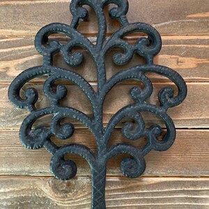 Puede incluir: Salvamanteles de hierro fundido negro con forma de árbol y detalles de volutas intrincadas. Tiene un lazo en la parte superior para colgar y un detalle en forma de diamante en la parte inferior. El fondo es una superficie de madera.