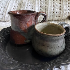 Puede incluir: Dos tazas de cerámica hechas a mano con asas. Una taza es de color marrón rojizo oscuro con detalles verdes y naranjas. La otra es una taza más pequeña, de color gris verdoso. Ambas están sobre una bandeja negra ornamentada, lo que sugiere un tema rústico o artesanal.