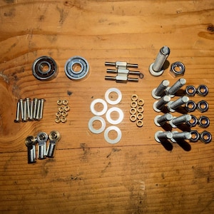 May include: An assortment of metal hardware components, including bearings, bolts, washers, and nuts, arranged on a wooden surface. The hardware is silver and gold in color, with various sizes and shapes.