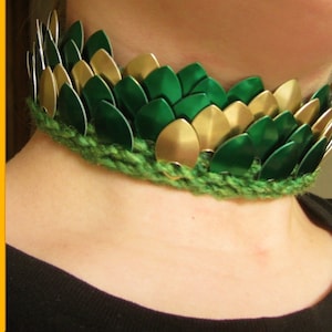 Puede incluir: Un collar gargantilla de malla de cadena verde y dorado con una banda de crochet verde. El collar está hecho de escamas de metal y se lleva alrededor del cuello.