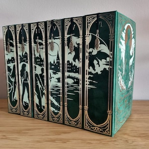 Op de afbeelding: Een set van zeven hardcover boeken in een decoratieve doos. De boeken zijn donkergroen met gouden accenten en bevatten ingewikkelde illustraties van figuren, gebouwen en wezens. Elk boek heeft een decoratief bladwijzer.