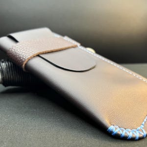 Puede incluir: Una funda de cuero marrón con un borde cosido azul y una correa de cuero marrón. La funda tiene una solapa curva y se combina con un lápiz óptico negro y gris. La funda está diseñada para guardar un lápiz óptico o un artículo similar.