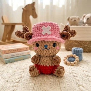 May include: A handmade crochet plush toy of a cartoon character, featuring a pink hat with a white cross, brown antlers, and red shorts. The plush is brown and tan with black eyes and a blue nose. The toy is sitting on a white, textured surface.