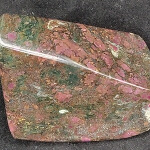 Puede incluir: Una gema rectangular pulida con esquinas redondeadas. La piedra presenta un patrón moteado de tonos rojo oscuro, verde y marrón. La superficie brillante refleja la luz, mostrando las variaciones naturales de color y textura.